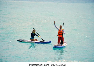 Young Man Dressed In Swimsuit And Lifevest Waving And Young Woman Paddling On Stand Up Paddle Board. Summer Outdoor Seaside Activities