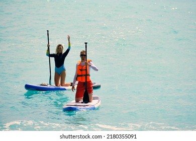 Young Man Dressed In Swimsuit And Lifevest Paddling And Young Woman Waving On Stand Up Paddle Board. Summer Outdoor Seaside Activities