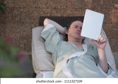 A Young Man In A Dressing Gown Lies On A Sofa Under A Canopy With A Tablet