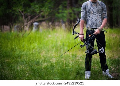 Young man dressing in jeans and a camo shirt looking down at a bow and arrow in his hands. He is holding a compound bow and stands in a mountain field.  - Powered by Shutterstock