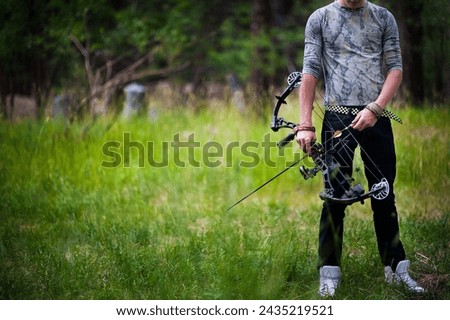 Young man dressing in jeans and a camo shirt looking down at a bow and arrow in his hands. He is holding a compound bow and stands in a mountain field. 