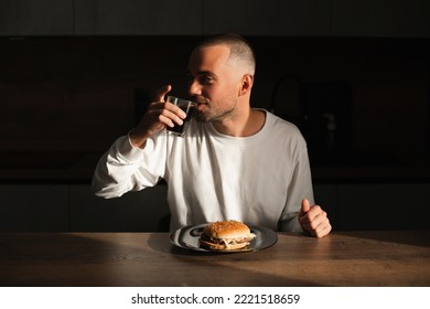 Young Man Drink Cola. Delicious Meal. Portrait Of Hungry Male With Cola And Burger. Smiling Casual Man Tasty Fast Food, Chewing Snack Sitting On Kitchen At House. Unhealthy Food.