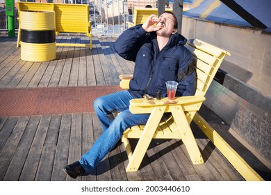 Young man drinking alcohol from little glass, head thrown back, sitting on wooden yellow armchair on sunny terrace in blue jeans and warm jacket.