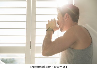 Young man drinking coffee and looking through sunlit bedroom window in morning