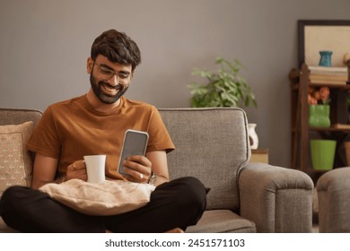 Young man drinking coffee and using mobile phone while sitting on sofa in living room - Powered by Shutterstock