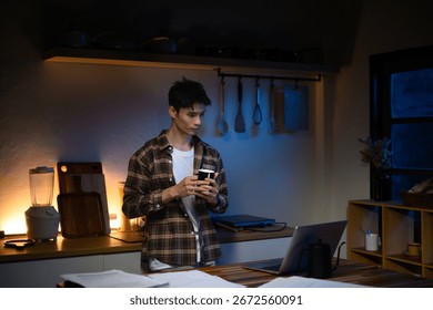 Young man drinking coffee while working late on a laptop in the kitchen