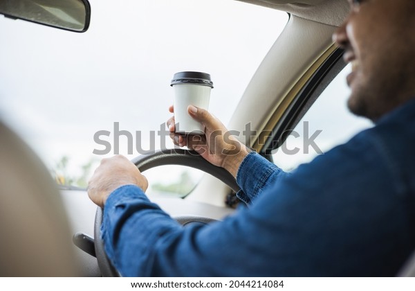 Young man drinking a cup of hot\
coffee while driving car to travel. Hands holding steering\
wheel.