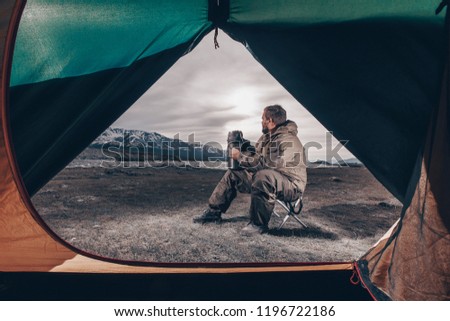 Young man drinking tea or coffee in mountains. Mountain camping concept. Traveler man with beard sit near the mountain lake . Man traveler hands holding cup of tea. the view from the tent