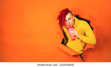 Young man drinking through soft drink straw in hole of orange background. Bright guy enjoying nonalcoholic beverage - Shutterstock ID 2134702635