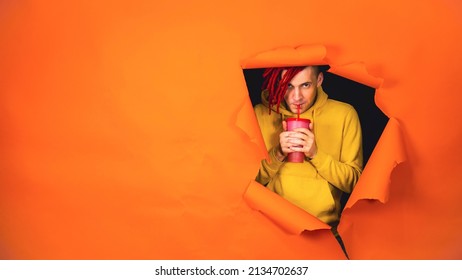 Young man drinking through soft drink straw in hole of orange background. Bright guy enjoying nonalcoholic beverage - Shutterstock ID 2134702637