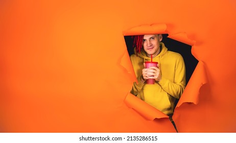 Young man drinking through soft drink straw in hole of orange background. Bright guy enjoying nonalcoholic beverage - Shutterstock ID 2135286515