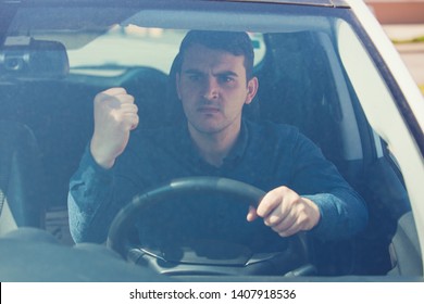 Young Man Driver Sitting In His Car Behind The Wheel Shaking His Fist Threatens Another Motorist. Bad And Aggressive Behavior On Road Concept.