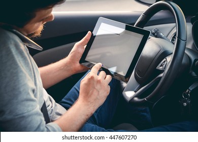 Young Man Driving A Car With A Tablet In Hand, Talking On Mobile Phone. Multitasking. Safe Driving Concept