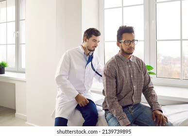 Young Man During Health Checkup At Doctor's Office. Physician At Modern Clinic Or Hospital Sitting On Medical Exam Couch And Using Stethoscope To Examine Respiration Or Heartbeat Of His Male Patient