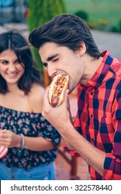 Young man eating an american hot dog and woman laughing on the background in a outdoors summer barbecue with his friends