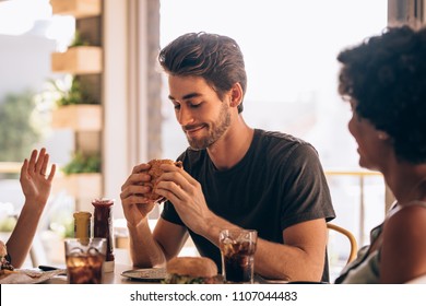 Young Man Eating Burger While Sitting With Female Friends At A Restaurant. Man Hanging Out At Cafe With Friends And Eating Burger.