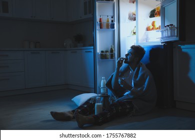 Young man eating cookies near open refrigerator in kitchen at night