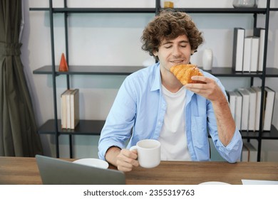 young man eating croissant while working on laptop computer at home - Powered by Shutterstock