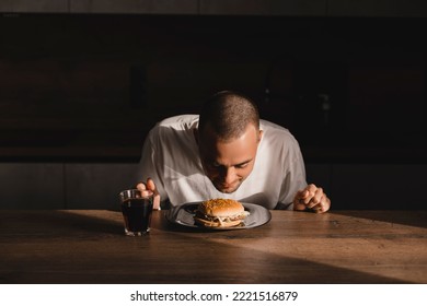 Young Man Eating A Hamburger. Delicious Meal. Portrait Of Hungry Male Model Smell Burger. Smiling Casual Man Holding And Smelling Tasty Fast Food, Chewing Snack With Closed Eyes Sitting On Kitchen.