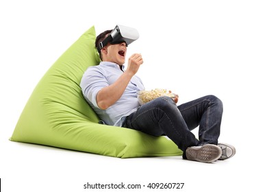 Young man eating popcorn and watching something on a VR goggles seated on a beanbag isolated on white background - Powered by Shutterstock