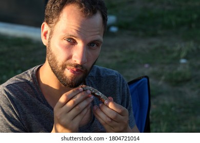 Young Man Eating Ribs At The Barbecue In A Park Sitting In A Tourist Chair