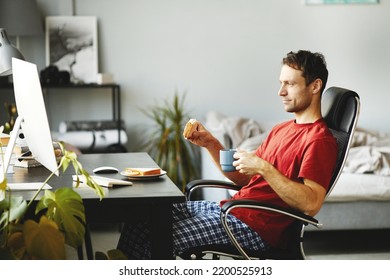 Young Man Eating Sandwich And Drinking Coffee While Sitting On Chair In Front Of Computer And Watching Video