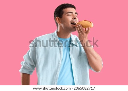 Young man eating tasty hot dog on pink background