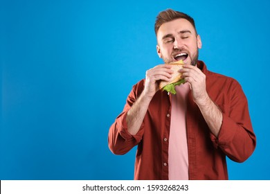 Young man eating tasty sandwich on light blue background. Space for text