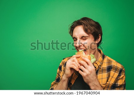 Young man eats burger with pleasure, isolated on green background