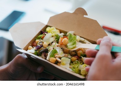 a young man eats a chickpea salad from a brown paper container, using a bamboo fork, sitting at his desk