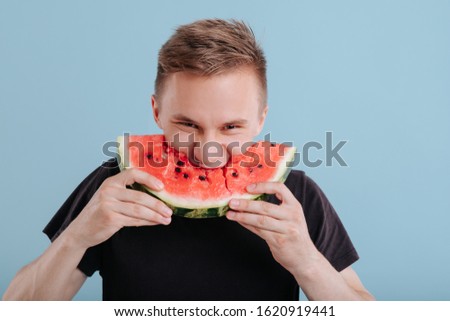 young man eats slice of watermelon, with lust, in studio, isolated on blue background, copy space
