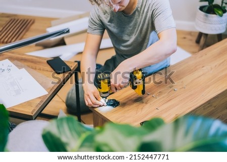 A young man with an electrical screwdriver assembles a tv stand console according to instructions in his new house. Man assembling furniture at home using a cordless screwdriver. Selective focus.