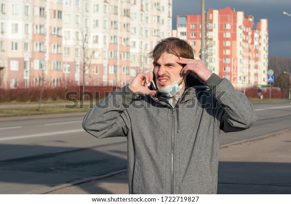 Young man emotionally talking on a cell phone\
in the street gesturing with his hands. On the face a protective\
mask from coronavirus. Protection against viruses transmitted by\
airborne droplets.