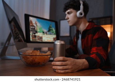 Young man with energy drink and headphones playing video game at wooden desk indoors, focus on can