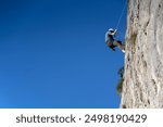 A young man is engaged in rock climbing, mountaineering in a beautiful mountainous area with red rocks, an athlete in a helmet descends from the top against the blue sky on a rope.