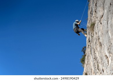 A young man is engaged in rock climbing, mountaineering in a beautiful mountainous area with red rocks, an athlete in a helmet descends from the top against the blue sky on a rope. - Powered by Shutterstock