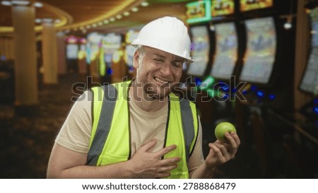 Young man engineer in hardhat and highvis vest clutch chest heartattack holding green apple in casino building; pain workplace hazard.