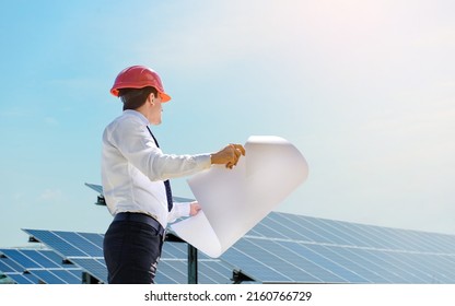 Young man engineer in white shirt and red safety helmet holding a project paper plan and verifying the photovoltaic panels at the solar power plant. Copy space for your text. - Shutterstock ID 2160766729