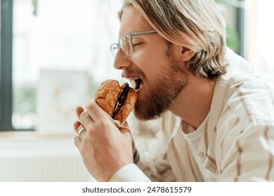 Young man enjoying a burger during a break from remote work in his cozy home office, savoring every bite with a satisfied smile - Powered by Shutterstock