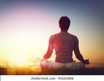 Young Man Enjoying Meditation And Yoga On Green Grass On Sunset Sky Background.