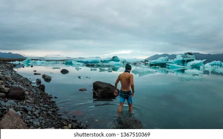 Young man enters the icy cold waters of Glacier lagoon. Man wearing only swimming shorts. Ice bergs drifting in the lagoon. Cold temperatures for ice swimming. Calm surface of the water
