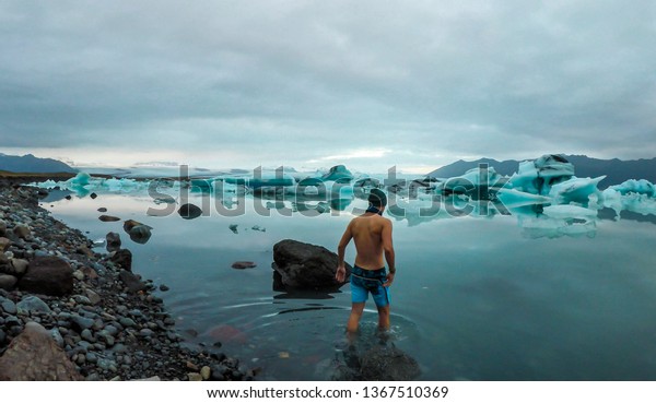 Young man enters the icy cold waters of Glacier\
lagoon. Man wearing only swimming shorts. Ice bergs drifting in the\
lagoon. Cold temperatures for ice swimming. Calm surface of the\
water