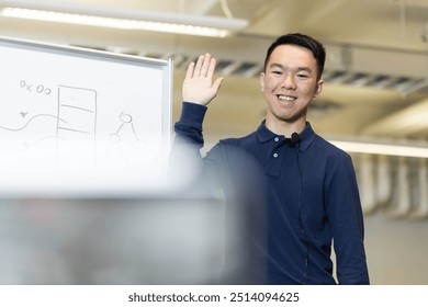 A young man is enthusiastically waving at the camera in front of a whiteboard while streaming a remote learning lesson to his students