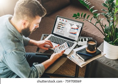 Young man, entrepreneur, freelancer sits at home on couch at coffee table, uses smartphone, working on laptop with graphs, charts, diagrams on screen.Online marketing,education, e-learning. Startup.