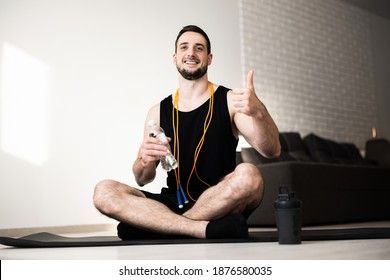 Young Man Exercising At Home. Guy Sits Alone On Black Yoga Map, Holds Water Bottle And Thumbs Up. Yellow Jumping Rope Around Neck. Athletist Rest And Relax. Small Break.