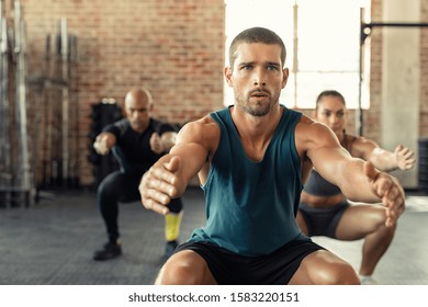 Young man exercising with squat in gym with people in background. Fit man exercising with stretched hands and squats at gym. Fitness class squatting togeher with outstretched during an exercise.