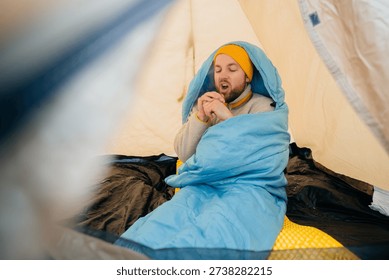 Young man experiencing cold while sleeping in a tent, rubbing hands together to generate warmth on winter camping trip