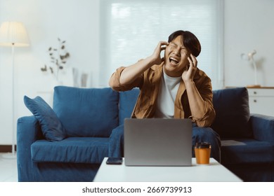 Young man experiencing a headache while working on a laptop at home, sitting on a sofa and dealing with stress and exhaustion