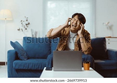 Young man experiencing a headache while working on a laptop at home, sitting on a sofa and dealing with stress and exhaustion