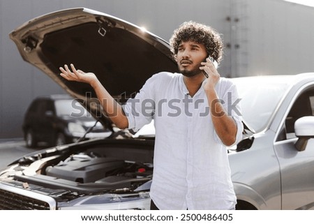 Young man is experiencing transportation difficulties, standing next to his broken down car with the hood raised while making a phone call for assistance